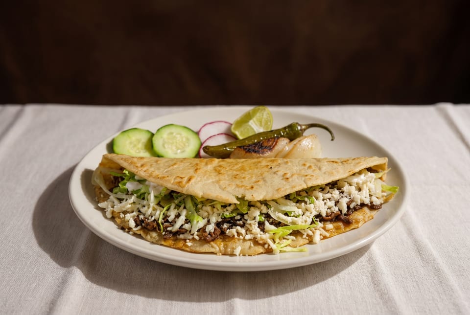 A large quesadilla filled with meat, lettuce, and cheese, served with cucumber, radish, and lime.